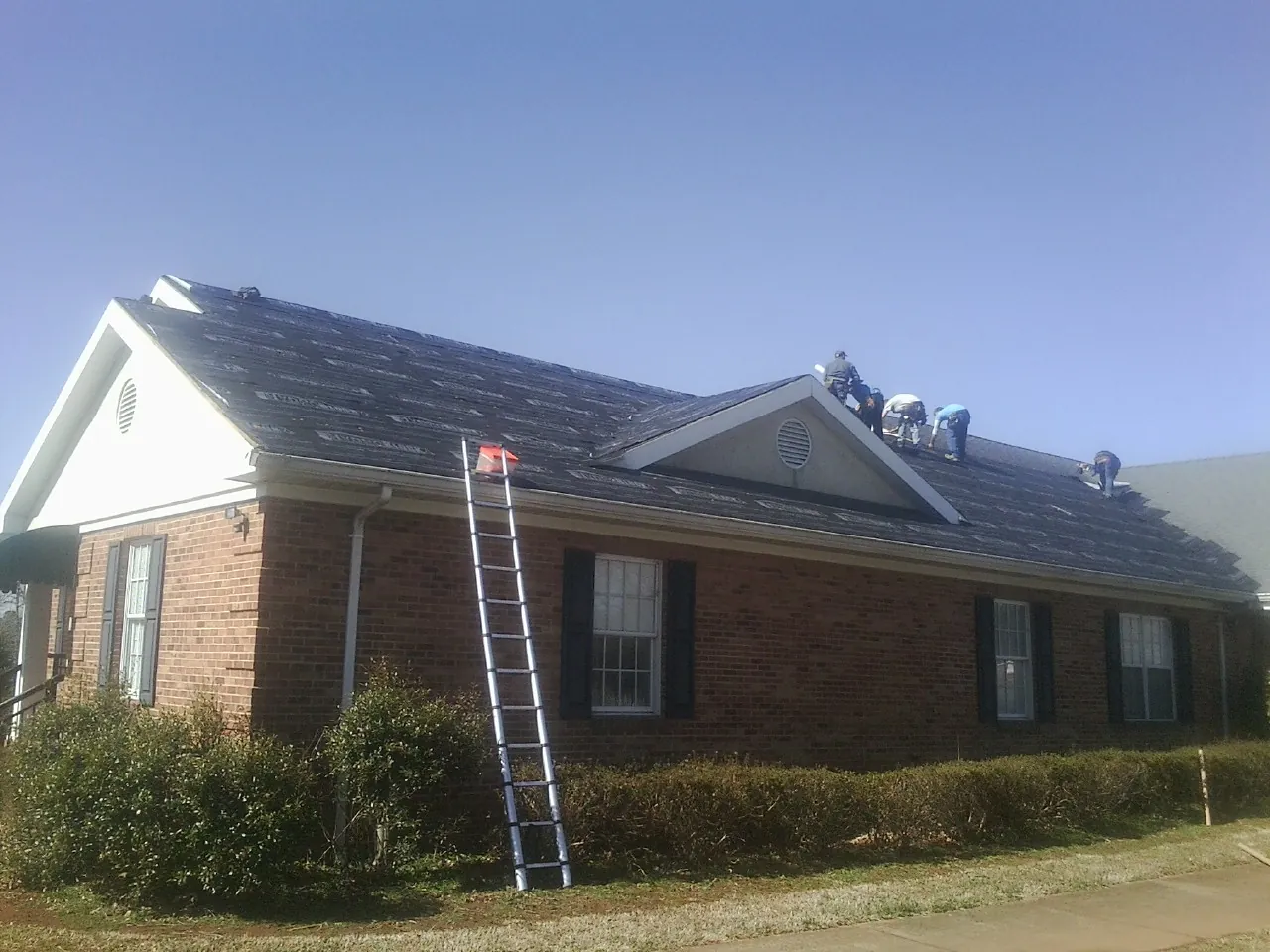 Crew tearing shingles off a brick ranch roof with a ladder set — Metal Roof Repair work by our crew in La Belle