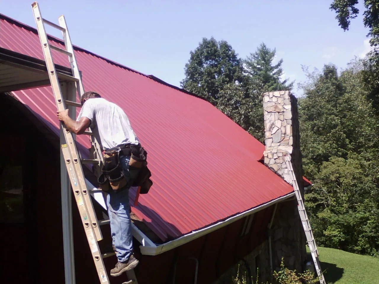 Roofer on a ladder beside a red metal roof and stone chimney — Soffit & Fascia Repair by Roofing Brothers in La Belle